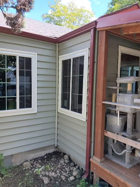 Corner of a beige house with two new windows and a red gutter line beside a covered porch